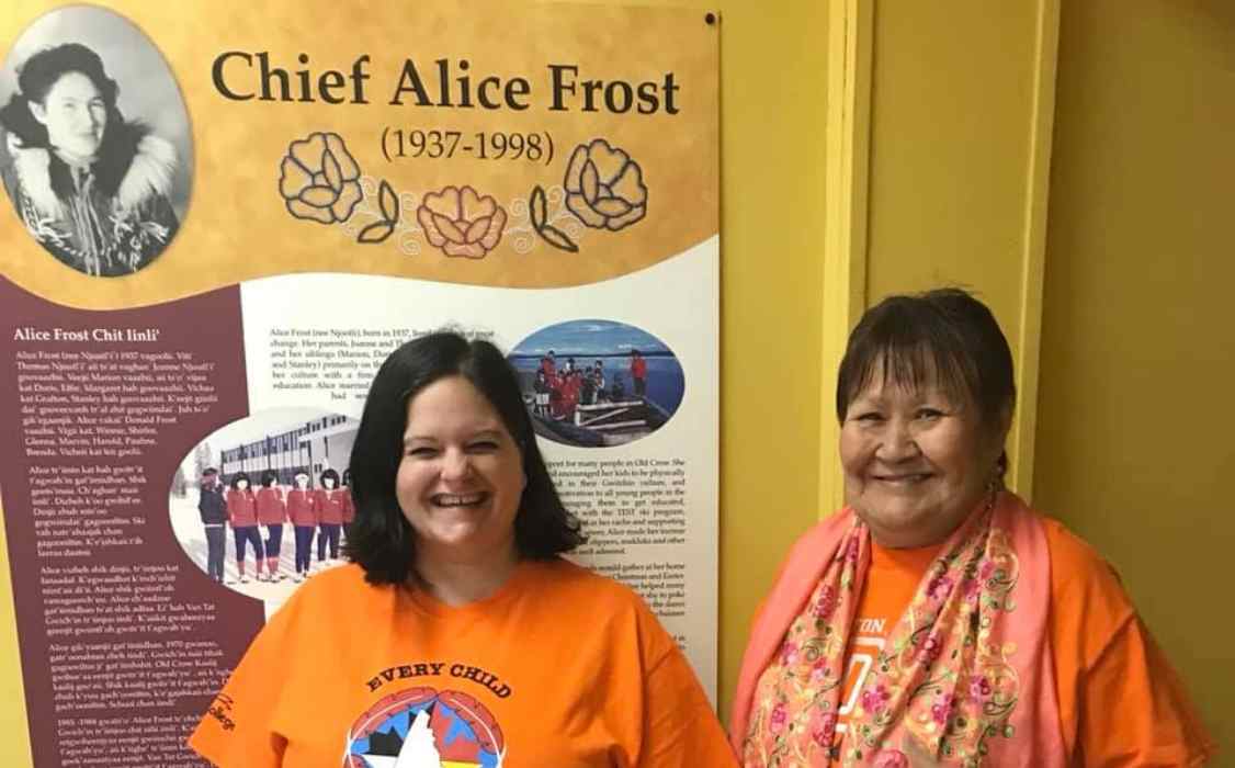 Two women wearing orange shirts and standing in front of a poster with a biography of Chief Alice Frost