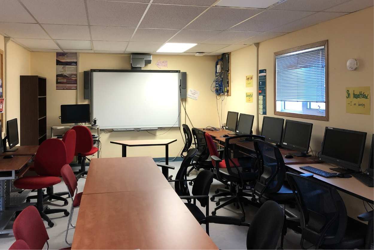 A computer lab with a series of PC computers along each wall with rolling chairs in front of them. A whiteboard is on the wall at the far end of the room.