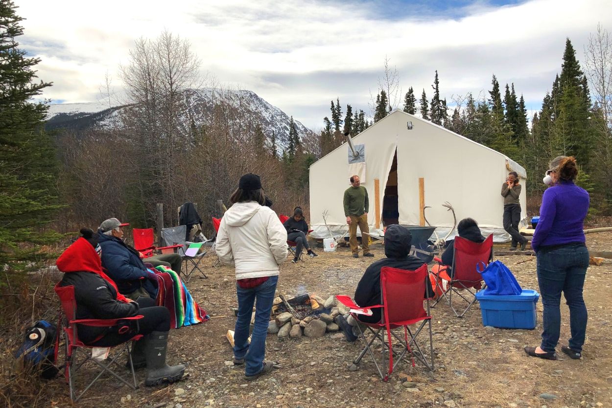 Students and elders around a fire, some sitting in camp chairs, next to a large wall tent.