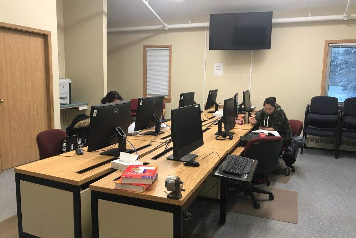 Two facing rows of desks with PC computers. Students sit on rolling chairs at the desks. A TV is mounted above the desks on the far wall.