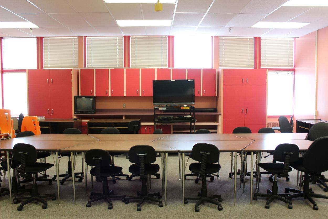 A row of trapezoid desks and chairs facing a TV screen. Behind the TV are orange-red book cases.