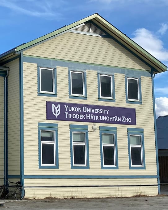 Pale yellow building with blue and green trim and two levels of windows. A purple sign above the first row of windows reads Yukon University, Tr'odëk Hätr'unohtän Zho