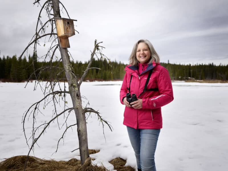 Katie Aitken posing outdoors holding binoculars next to a tree with a birdhouse