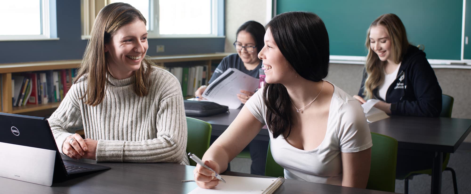 Four smiling students sitting at two rows of tables in a classroom. One student is writing in a notebook, one has a laptop computer, and the other two are reading.