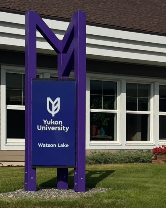 A purple sign for Yukon University Watson Lake outdoors in front of a row of windows