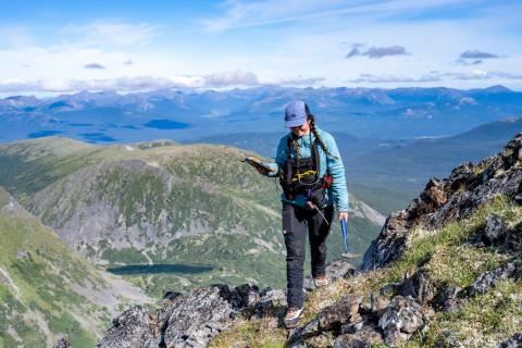 woman walks along mountain ridge holding pick with a clear mountain view behind