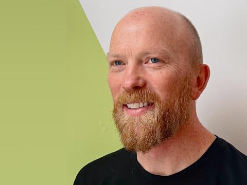 A smiling bearded man with a shaved head in front of a green and white wall