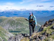 woman walks along mountain ridge holding pick with a clear mountain view behind