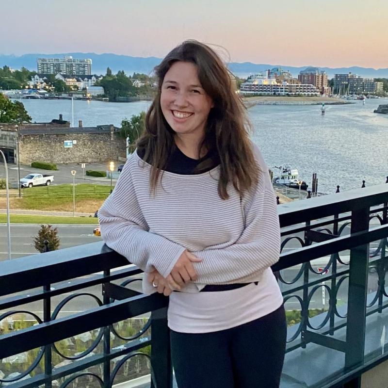 A woman with long brown hair standing on a patio with a view of a cruise ship in harbour behind her