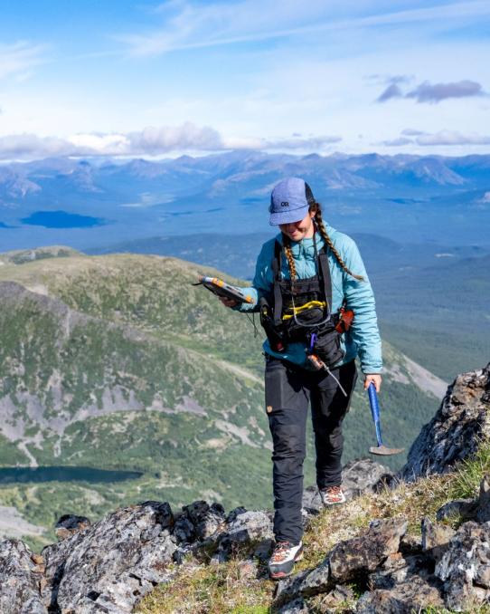 woman walks along mountain ridge holding pick with a clear mountain view behind