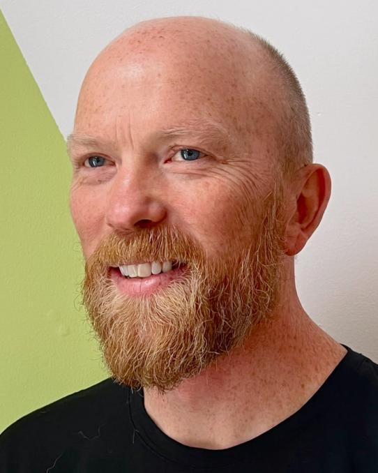 A smiling bearded man with a shaved head in front of a green and white wall